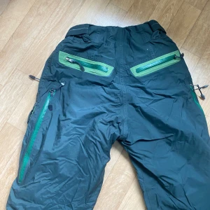 Svarta täckbyxor från Ace Storm Fit M - Svarta täckbyxor från Ace Storm Fit NIKE med gröna detaljer och dragkedjor. Byxorna har flera fickor med dragkedja och förstärkta partier vid bensluten. Insidan är fodrad med grönt tyg för extra värme. Perfekta för vinteraktiviteter och snö.