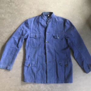 Blå overshirt med ståkrage - Snygg blå overshirt med ståkrage och fyra stora fickor framtill, alla med knappstängning. Jackan har raka linjer, knappar längs fronten och är tillverkad i ett slitstarkt bomullstyg. Perfekt för dig som gillar minimalistisk och funktionell stil.