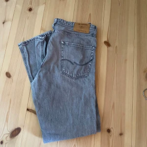 Grå jeans - Säljer ett par Jack and Jones jeans i grått med straight fit. De har en snygg femficksdesign, ikonisk läderpatch bak och coola kontrastsömmar på bakfickan. Perfekta för dig som gillar tidlös stil och vill ha ett par jeans som funkar till allt. Mycket fint skick.