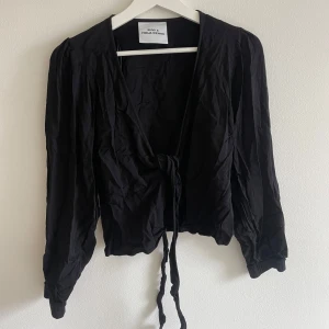 Svart omlottopp från Envii x Freja Wewer - Snygg svart omlottopp med knyt framtill från Envii x Freja Wewer. Toppen har långa ärmar med lätt puff och elastiska muddar. Croppad passform och mjukt, tunt material som känns skönt mot huden. Perfekt för dig som gillar stilrena och trendiga plagg.