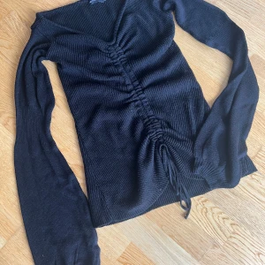 Svart ribbad tröja PrettyLittleThing - Svart stickad offshoulder-tröja från PrettyLittleThing med ribbad struktur och snygg snörning framtill. V-ringad och långärmad modell som sitter tajt och har en croppad känsla.