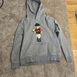 Grå hoodie Polo Bear Ralph Lauren - Säljer en grå hoodie från Polo Ralph Lauren med ikoniska Polo Bear-trycket på bröstet. Tröjan har huva och långa ärmar, perfekt för en chill och sportig look. Tillverkad i mjuk bomullsmix som är skön att ha på sig hela dagen.