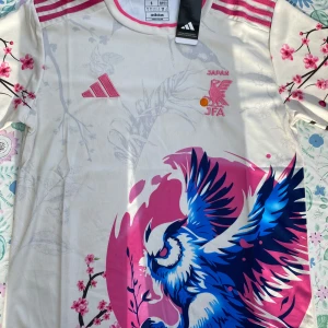 Japan Adidas fotbollströja S - Unik Japan fotbollströja från Adidas i slim fit med korta ärmar. Tröjan är vit med rosa detaljer, har ett stort blått och rosa fågeltryck framtill och körsbärsblommor på ärmar och rygg. Materialet är lätt polyester med AEROREADY-teknologi för bra ventilation.