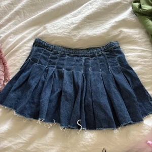 Mörkblå plisserad jeanskjol - Säljer en mörkblå plisserad jeanskjol med rå kant och fransig nederdel. Kjolen har en klassisk midjehöjd och är kort i modellen, perfekt för dig som gillar en edgy och trendig look. Materialet är robust denim och kjolen har snygga sömdetaljer.