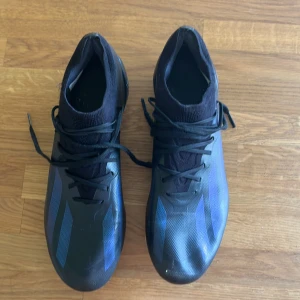 Adidas Crazyfast svarta fotbollsskor - Säljer ett par Adidas Crazyfast fotbollsskor i svart med blå detaljer och klassiska tre ränder på sidan. Skorna har snörning, låg profil och platt sula med dobbar för gräsplan. Ovandelen är i syntetmaterial och designen är riktigt sportig och clean.