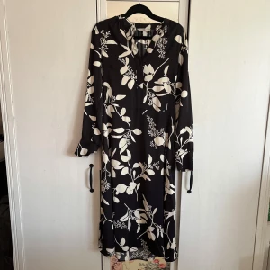 Svart blommig långklänning - En svart långklänning från H&M i strl S med vitt blommigt mönster. Klänningen har långa ärmar med en knapp vid ärmslut, v-ringad hals och ett bälte i midjan som är avtagbart. Har även satt på en nål som går att ta bort ifall det önskas!💞