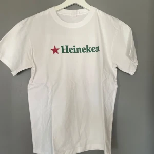 Vit t-shirt med Heineken-tryck - Snygg vit t-shirt med det ikoniska Heineken-trycket i grönt och en röd stjärna. Perfekt för en avslappnad stil eller för att visa din kärlek till Heineken. T-shirten har en klassisk passform och korta ärmar.