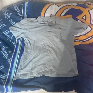 Ljusblå pikétröja med broderad logga - Säljer en ljusblå pikétröja med krage och knappar. Tröjan har en liten broderad logga på bröstet. Perfekt för en avslappnad stil.