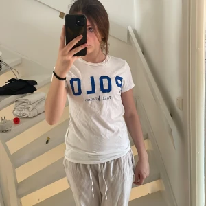 Vit t-shirt från Ralph Lauren - Säljer en klassisk vit t-shirt från Ralph Lauren med blå text 'POLO' på framsidan. T-shirten är kortärmad och har en normal passform. Perfekt för en avslappnad stil. 🕶️