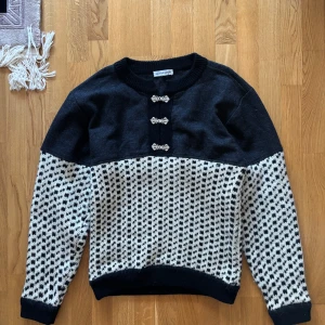 Svart och vit mönstrad ulltröja från Charade - Säljer en snygg svart och vit tröja från Charade med ett unikt mönster och dekorativa spännen framtill. Tröjan har långa ärmar och ribbade kanter vid hals, ärmslut och nederkant. Perfekt för kyligare dagar!