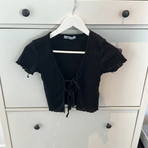 Svart ribbad bolerotopp från Gina Tricot - Snygg svart ribbad bolerotopp från Gina Tricot med korta ärmar och knytning framtill. Perfekt för att lägga till en stilren touch till din outfit.