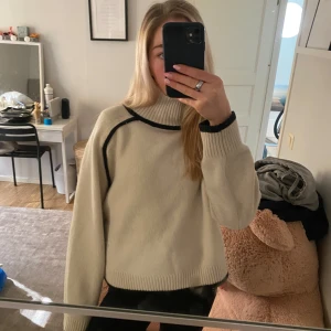 Beige polotröja med svarta detaljer - Säljer en snygg beige polotröja med svarta kontrastlinjer. Tröjan har en hög krage och långa ärmar, perfekt för kyligare dagar. Den ribbade kanten ger en stilren look. Passar bra till både jeans och kjol!
