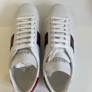 Vita sneakers från Gucci - Säljer ett par helt oanvända och nya vita sneakers från Gucci med klassiska gröna och röda ränder på sidan. Skorna har en broderad bi-detalj och snörning framtill. Baktill finns en kontrasterande häl i grönt och rött. 