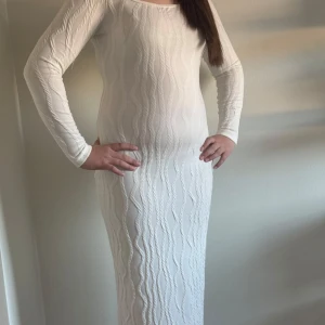 Vit långklänning med vågigt mönster - Elegant vit långklänning med ett subtilt vågigt mönster. Klänningen har långa ärmar och en tight passform som framhäver figuren. Perfekt för en stilren look.