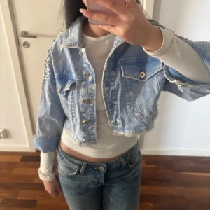 Jeansjacka med pärldetaljer från Qishe - Snygg croppad jeansjacka från Qishe med pärldetaljer på axlarna. Jackan har en ljusblå tvätt och knappar framtill. Perfekt för att ge din outfit en cool touch. Vintage!