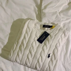 Vit kabelstickad tröja från Polo Ralph Lauren - Säljer en vit sweater från Polo Ralph Lauren. Tröjan har en klassisk rund hals och är prydd med den ikoniska loggan på bröstet. Perfekt för en elegant och avslappnad look.