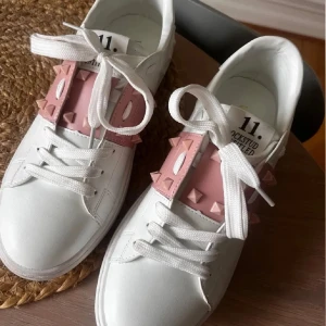 Valentino sneakers med rosa detaljer - Snygga vita sneakers från Valentino med rosa detaljer och nitar. Skorna har snörning och en rund tå, vilket ger en stilren look. Perfekta för att addera en touch av lyx till din outfit.💕