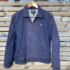 Ralph lauren windbraker marinblå XS -  En riktigt skön Ralph Lauren windbreaker i marinblå färg och storlek XS är en superfin jacka! Den är lätt och skyddar mot vinden, perfekt för att vara ute! 🌬️ Den har en snygg design och elastiska ärmslut som gör att den sitter bra.   Marinblått är en klassisk färg som passar till allt, så du kan enkelt matcha den med olika kläder! Det är en toppenjacka för blåsiga väder, en toppen jacka för  utflykter eller bara för att hänga med kompisar🙌 hör av dig vid frågor eller funderingar! 