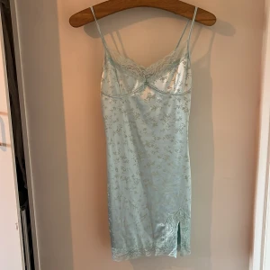 Ljusblå klänning från Motel rocks  - Säljer en elegant ljusblå klänning från Motel med spetsdetaljer och blommigt mönster. Klänningen har smala axelband och en liten slits framtill. Perfekt för en somrig look!