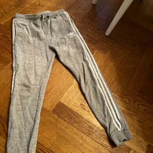 Grå mjukisbyxor från Adidas - Säljer ett par grå mjukisbyxor från Adidas med klassiska vita ränder längs sidorna. Byxorna har en bekväm resårmidja med snörning och är perfekta för en avslappnad stil. Tillverkade i mjukt bomullsmaterial för extra komfort.