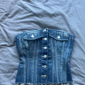Jeans-korsett från Zara - Snygg jeans-korsett från Zara med knappar framtill och fransig nederkant. Den har en cool denim-look och är perfekt för att ge en edgy touch till din outfit. Baksidan har en elastisk del för bättre passform.