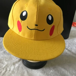 Barn Pikachu keps - Säljer en supercool gul keps med Pikachu-motiv. Kepsen har justerbar passform och är perfekt för alla Pokémon-fans. Den har Pikachus ansikte fram och är gjord i bomull.  Ny 