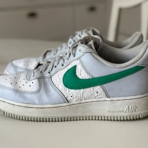 Nike Air Force 1  - Snygga vita Nike Air Force 1 'Embossed Hoops - Bone Green' med en grön swoosh och perforerade detaljer på ovandelen. Skorna har klassisk snörning och en bekväm sula med AIR-dämpning. Perfekta för en stilren look.