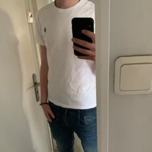 Vit t-shirt från Ralph Lauren - Säljer en klassisk vit t-shirt från Ralph Lauren med en liten broderad logga på bröstet. T-shirten har en rund hals och korta ärmar, perfekt för en stilren look. Tillverkad i bomull för en bekväm passform. Passar utmärkt till jeans eller shorts.