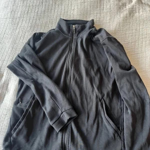 Svart hugo boss fullzip - Säljer en svart hugo boss tröja med dragkedja och hög krage. Jackan har långa ärmar och två fickor framtill. Perfekt för kyliga dagar.