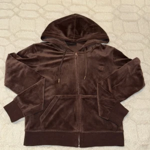 Brun hoodie från Juicy Couture - Säljer en brun hoodie från Juicy Couture i mjukt material. Den har dragkedja framtill och en praktisk huva med snörning. Perfekt för en avslappnad stil.