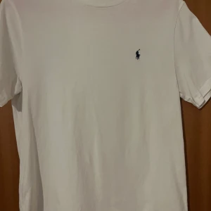 Vit T-Shirt Polo Ralph Lauren  - Säljer en klassisk vit t-shirt från Polo Ralph Lauren med den ikoniska loggan broderad i blått på bröstet. Perfekt för en stilren och avslappnad look. Tillverkad i mjuk bomull för extra komfort. Storlek L Passar 165-180