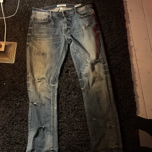 Blå jeans med slitningar - Säljer ett par blå jeans med coola slitningar och en klassisk femficksdesign. De har en knappgylf och är i en straight passform. Perfekta för en avslappnad stil.