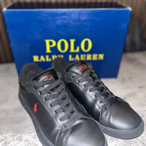 Svarta sneakers från Polo Ralph Lauren - Snygga svarta sneakers från Polo Ralph Lauren med röd logga på sidan. Skorna har snörning och en rund tå. Perfekta för en stilren look.