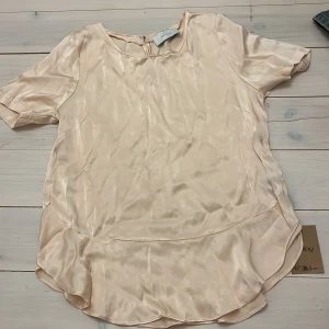 Beige satinblus från Neo Noir - Säljer en  beige/rosa satinblus från Neo Noir. Blusen har korta ärmar och en rund halsringning. Den har en lätt glansig färg och en längre bakdel för en stilfull look. Köpt på Sellpy och aldrig använd 