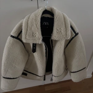 Vit teddyjacka från Zara - Säljer en snygg vit teddyjacka från Zara med svarta detaljer. Jackan har en oversized passform och dragkedja framtill. Perfekt för kyliga dagar med sin mysiga och varma design. Aldrig använd, bara testad! 