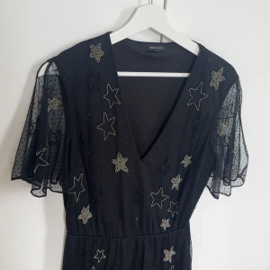 Svart klänning med stjärnor från River Island - Snygg svart klänning från River Island med glittriga stjärnor och v-ringning. Klänningen har korta, transparenta ärmar med ett lätt puffigt utseende. Perfekt för en kväll ute eller en speciell tillställning.