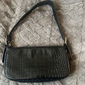 Svart axelväska med krokodilmönster - Snygg svart axelväska med krokodilmönster och dragkedja. Perfekt för att ge en stilren touch till din outfit. Väskan har en justerbar rem och är tillverkad i syntetmaterial.❤️🥰