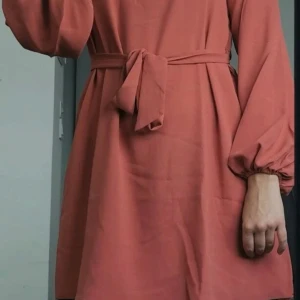 Korallfärgad klänning med knytband - Snygg korallfärgad klänning med långa ärmar och ett knytband i midjan. Perfekt för en stilren look. Klänningen har en lös passform och är tillverkad i ett mjukt material.