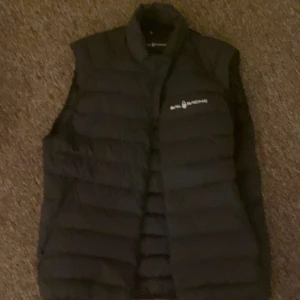 Svart dunväst från Sail Racing - Snygg svart dunväst från Sail Racing med dragkedja och logotyp på bröstet. Västen är vindtät och andas bra, perfekt för kyliga dagar. Den har en stilren design och är idealisk för både vardag och utomhusaktiviteter. Priset går diskutera 