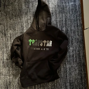 Trap Star Hoodie - Säljer en svart hoodie med tryck på framsidan i grönt och vitt. Den har en klassisk design med en stor ficka framtill och en bekväm huva. 