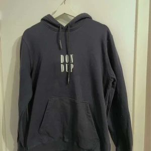 Mörkblå hoodie från Dondup - Säljer en snygg mörkblå hoodie från Dondup med vit logga på bröstet. Tillverkad i 100% bomull, vilket gör den både bekväm och hållbar. Hoodien har en klassisk känguruficka och justerbar dragsko i huvan. Perfekt för en avslappnad stil.