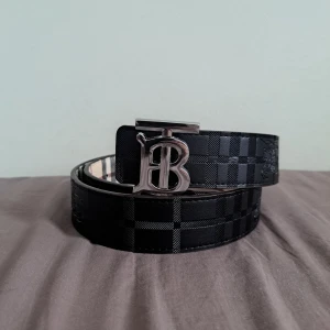 Burberry bälte - Snyggt vändbart bälte. Ena sidan har ett klassiskt svart rutmönster medan den andra sidan har ett beige och rödaktigt rutmönster. Perfekt för att variera din stil! Storlek: 105CM