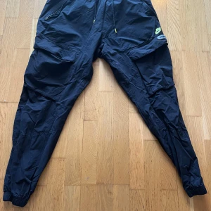 Svarta träningsbyxor från Nike - Säljer ett par svarta träningsbyxor från Nike med elastisk midja och flera praktiska fickor. Byxorna har en avslappnad passform och är perfekta för sportiga aktiviteter. Nike-loggan syns tydligt på sidan.