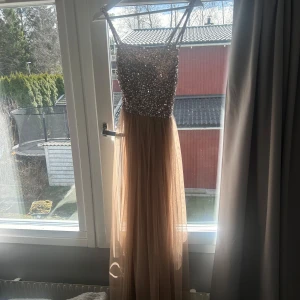 Champagnefärgad långklänning med paljetter - Elegant långklänning i champagnefärg med glittrande paljetter upptill och ett luftigt, flödande kjolparti. Klänningen har smala axelband som korsas i ryggen för en stilfull look. Finns en fläck på underkjolen som man ser på bilden.🩷