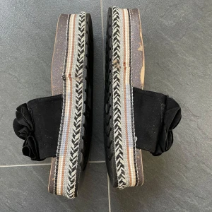 Svarta sandaler med fina detaljer  - Snygga svarta sandaler  med en unik volangdetalj på ovansidan. Sandalerna har en platåsula med randigt mönster i svart och vitt, vilket ger dem en trendig look. Perfekta för sommarens alla tillfällen. Använda vid två kortare tillfällen 
