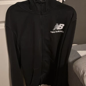 Svart hoodie från New Balance - Säljer en svart hoodie från New Balance med dragkedja framtill och logga på bröstet. Perfekt för en sportig look eller avslappnade dagar. Den har långa ärmar och en bekväm passform.
