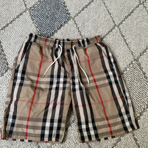 Beige burberry shorts  - Snygga beige burberry shorts med klassiskt rutig mönster i svart, vitt och rött. De har elastisk midja med vit snörning och är knälånga. Perfekta för sommaren och har en avslappnad passform.