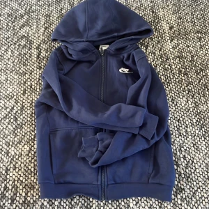 Mörkblå hoodie från Nike - Säljer en mörkblå hoodie från Nike med dragkedja och huva. Tröjan har två fickor framtill och en vit Nike-logga på bröstet. Perfekt för en sportig och avslappnad stil.
