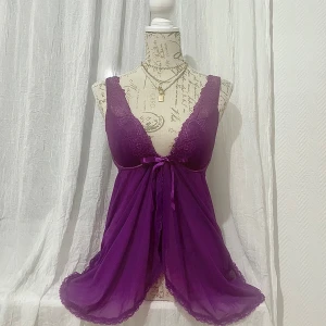 Lila negligé från Valege - Säljer en elegant lila negligé från Valege. Den har en vacker spetsdetalj upptill och ett knytband i midjan. Den är ärmlös och har en transparent design som ger en lyxig känsla. STORLEK 75D som passar byst 94-97cm. Korrekt färg på första bild.