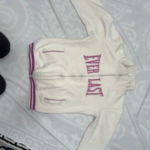 Vit ziptröja från Everlast - Vit ziptröja från Everlast med lila detaljer och stora bokstäver på bröstet. Tröjan har två fickor framtill, ribbade muddar och dragkedja hela vägen upp. Perfekt för en sportig och avslappnad stil.